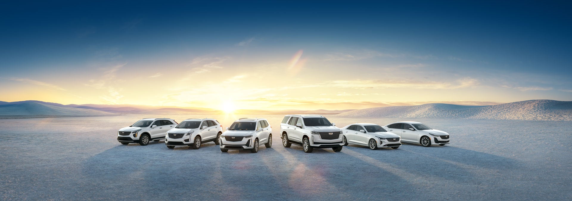 Several Cadillac vehicles parked on a flat surface with the sun rising in the background, casting a warm glow.